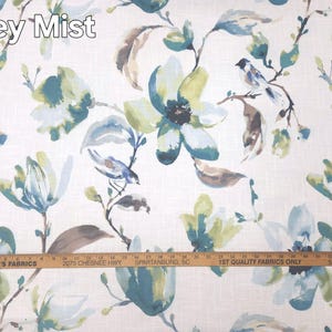 May include: A white fabric featuring a watercolor-style floral and bird pattern in shades of teal, blue, and muted green, labeled Zoey Mist. A wooden yardstick is positioned across the bottom, showing measurements in inches up to 36 inches.
