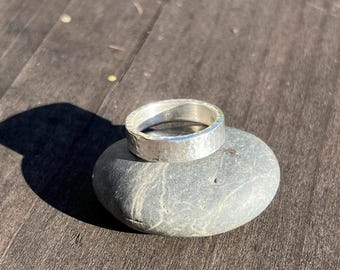 May include: A silver ring with a hammered texture rests on a smooth, gray stone. The ring has a wide band and a slightly irregular surface, reflecting the sunlight. The stone is oval-shaped, and the background is a dark wooden surface.