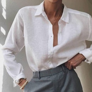 May include: A white button-down shirt with long sleeves and a collar, paired with gray trousers. The shirt is unbuttoned at the top, revealing a glimpse of the chest. A gold necklace and bracelets are visible.