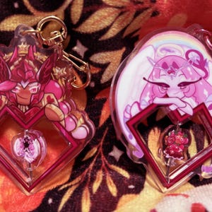 May include: Two acrylic keychains featuring cartoon characters. One keychain has a red and pink character with a crown, the other has a pink character with white wings and a rainbow. Both have a heart-shaped frame.