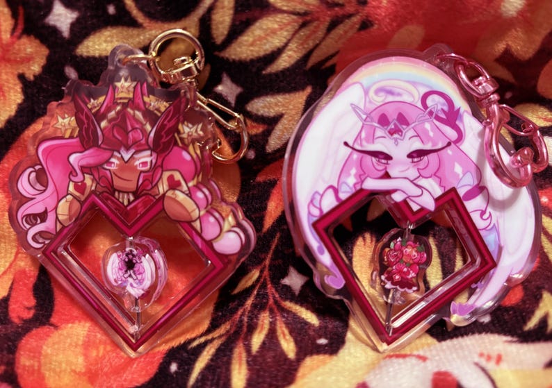 May include: Two acrylic keychains featuring cartoon characters. One keychain has a red and pink character with a crown, the other has a pink character with white wings and a rainbow. Both have a heart-shaped frame.