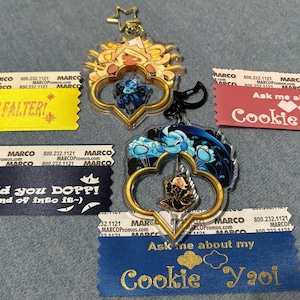 May include: Two clear acrylic keychains with gold-colored frames and character art, plus four ribbons with text. The keychains have a star and heart design. The ribbons say "I WILL NOT FALTER!", "What'd you DOPPI!", "Ask me about my Cookie Yuri", and "Ask me about my Cookie Yaoi".