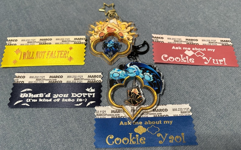 May include: Two clear acrylic keychains with gold-colored frames and character art, plus four ribbons with text. The keychains have a star and heart design. The ribbons say "I WILL NOT FALTER!", "What'd you DOPPI!", "Ask me about my Cookie Yuri", and "Ask me about my Cookie Yaoi".