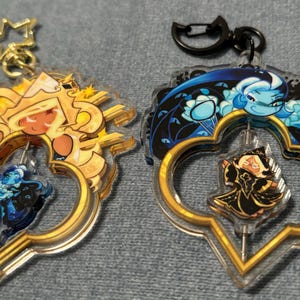 May include: Two clear acrylic keychains with gold-colored accents. One features a golden character with a blue figure inside, the other has a blue character with a black figure inside. Both have a star or a black clasp.
