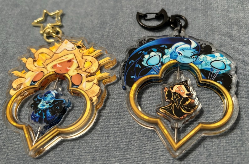 May include: Two clear acrylic keychains with gold-colored accents. One features a golden character with a blue figure inside, the other has a blue character with a black figure inside. Both have a star or a black clasp.