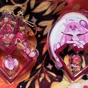 May include: Two acrylic keychains featuring anime-style characters. One keychain has a pink and red character with a crown and armor. The other has a pink character with white wings and a rainbow. Both have a heart-shaped frame.