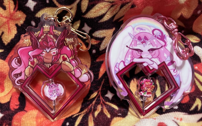 May include: Two acrylic keychains featuring anime-style characters. One keychain has a pink and red character with a crown and armor. The other has a pink character with white wings and a rainbow. Both have a heart-shaped frame.