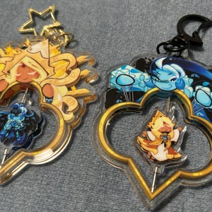 May include: Two acrylic keychains with gold-colored frames and character designs. One features a yellow and orange character, the other a blue and white character. Both have a gold star-shaped keychain attachment.