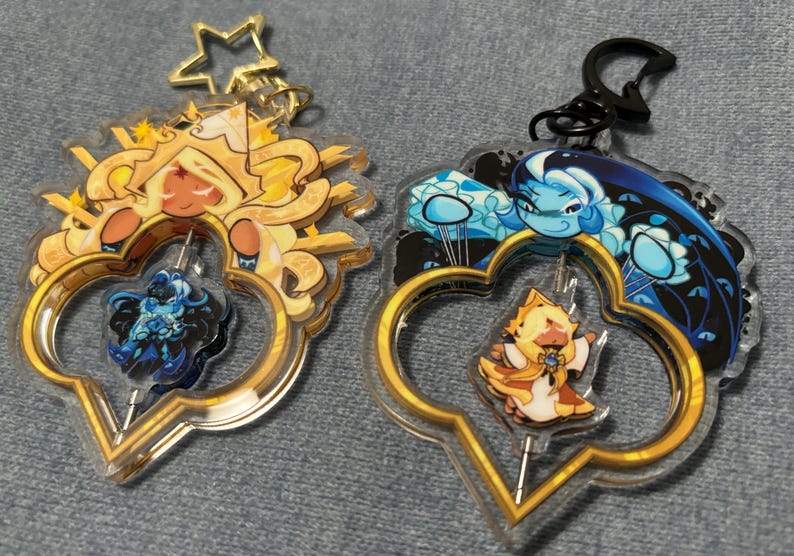May include: Two acrylic keychains with gold-colored frames and character designs. One features a yellow and orange character, the other a blue and white character. Both have a gold star-shaped keychain attachment.