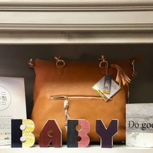 May include: A brown leather tote bag with gold hardware and a tassel. The bag is sitting on a shelf with wooden letters spelling out "BABY" and a sign that says "Do good."
