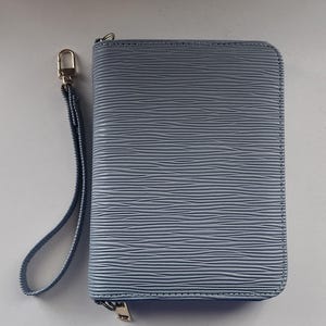 May include: A light blue, rectangular wallet with a textured, horizontal striped pattern. The wallet has a zipper closure and a wrist strap with a gold-colored clasp. The wallet is approximately 8 inches long and 4 inches wide.