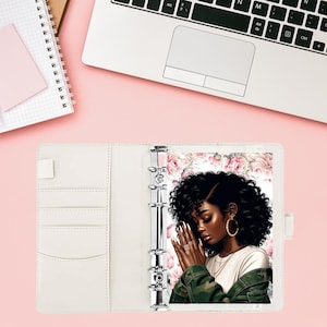 May include: A white planner with a floral design and a black and white illustration of a woman with her hands clasped in prayer. The planner has a ring binder system and a pocket on the front cover.