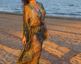 May include: A flowing, patterned kaftan dress in shades of brown, green, and blue, with a V-neckline and embellished trim. The dress is worn on a sandy beach with the ocean in the background. The fabric appears lightweight and sheer.