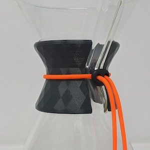 Puede incluir: Una jarra de café de vidrio transparente con una tapa de plástico negro y gris y un cordón de ajuste naranja brillante. La jarra está sobre una superficie negra y peluda.
