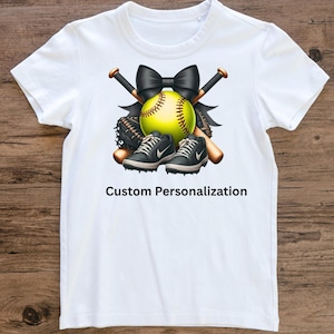 May include: White t-shirt featuring a softball design. The design includes a yellow softball, black bow, crossed baseball bats, a baseball glove, and black and gray sneakers. The text "Custom Personalization" is printed below the design.