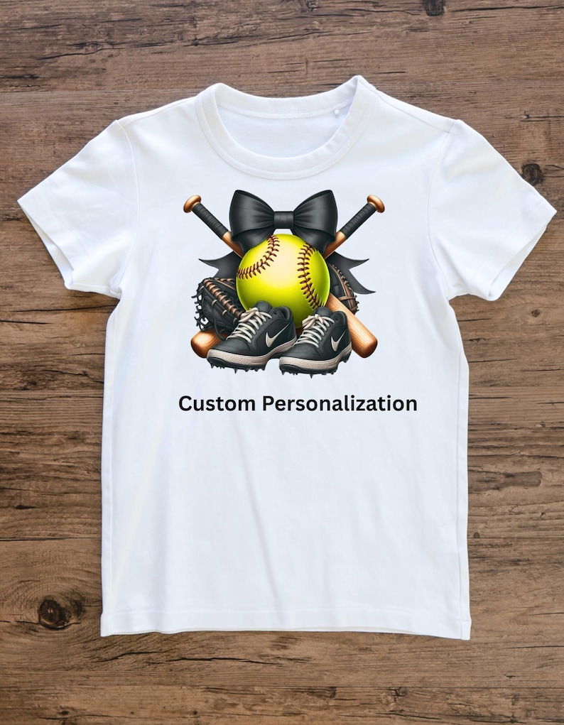 May include: White t-shirt featuring a softball design. The design includes a yellow softball, black bow, crossed baseball bats, a baseball glove, and black and gray sneakers. The text "Custom Personalization" is printed below the design.