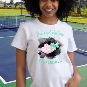 May include: White t-shirt with a graphic of a pink and white pickleball paddle with a green bow. The text "Just a girl who loves pickleball" is above the paddle.