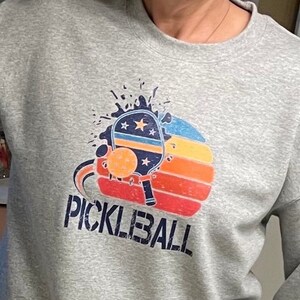 Puede incluir: Sudadera gris con un gr&aacute;fico de una pala de pickleball y una pelota en un dise&ntilde;o de puesta de sol. El texto "PICKLEBALL" est&aacute; impreso debajo del gr&aacute;fico.