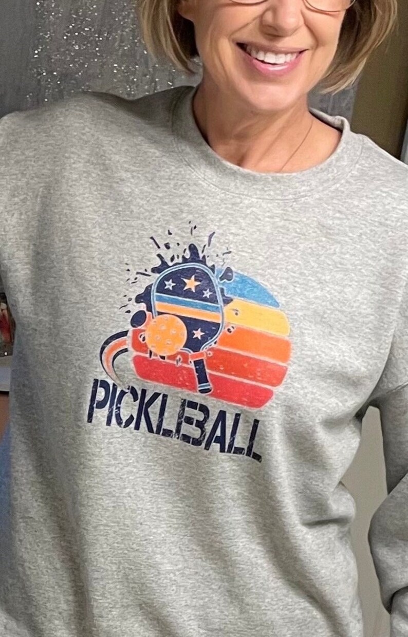 Puede incluir: Sudadera gris con un gr&aacute;fico de una pala de pickleball y una pelota en un dise&ntilde;o de puesta de sol. El texto "PICKLEBALL" est&aacute; impreso debajo del gr&aacute;fico.