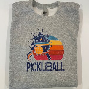 Puede incluir: Una sudadera gris con un gr&aacute;fico de estilo vintage de una pala de pickleball y una pelota con la palabra "PICKLEBALL" impresa debajo. El gr&aacute;fico presenta una puesta de sol con rayas rojas, naranjas y amarillas.