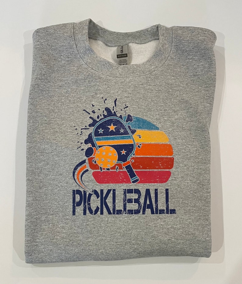 Puede incluir: Una sudadera gris con un gr&aacute;fico de estilo vintage de una pala de pickleball y una pelota con la palabra "PICKLEBALL" impresa debajo. El gr&aacute;fico presenta una puesta de sol con rayas rojas, naranjas y amarillas.
