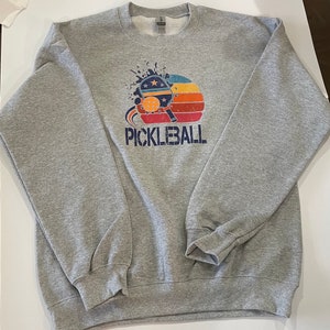 Puede incluir: Una sudadera gris con un gr&aacute;fico de estilo vintage de una pala de pickleball y una pelota con una puesta de sol detr&aacute;s. El texto "PICKLEBALL" est&aacute; impreso debajo del gr&aacute;fico.