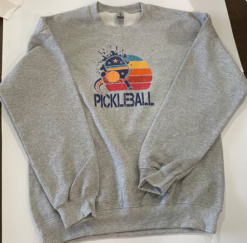 Puede incluir: Una sudadera gris con un gr&aacute;fico de estilo vintage de una pala de pickleball y una pelota con una puesta de sol detr&aacute;s. El texto "PICKLEBALL" est&aacute; impreso debajo del gr&aacute;fico.