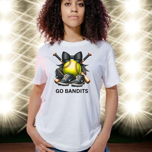 May include: White t-shirt featuring a softball design with a yellow softball, black bow, bats, and sneakers. The text "GO BANDITS" is printed below the design. The shirt is worn by a person.