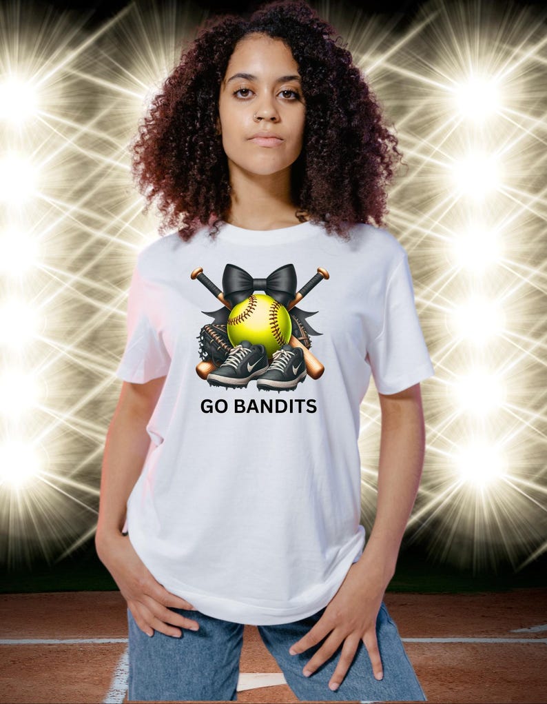 May include: White t-shirt featuring a softball design with a yellow softball, black bow, bats, and sneakers. The text "GO BANDITS" is printed below the design. The shirt is worn by a person.