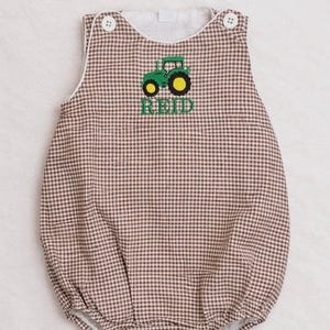 May include: Brown and white gingham baby romper with a green tractor applique and the name "REID" embroidered in green. The romper has white trim, button closures, and a rounded neckline. A cute outfit for a baby.