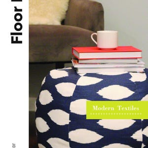 Puede incluir: Un puf de suelo azul y blanco con estampado con el texto "Modern Textiles" en una etiqueta verde. El puf está sobre un suelo de madera junto a una silla con una manta de piel de oveja.