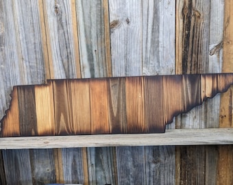 May include: A wooden state of Tennessee outline, burned with a dark brown finish. The wood is arranged in vertical planks, creating a rustic look.