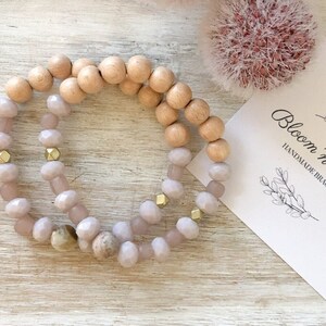 May include: Two bracelets, one with light pink and gold beads and the other with natural wood beads. The bracelets are stacked on top of each other on a light wood surface. The text "Bloom 'n Beady Handmade Bracelets" is visible on a white card.