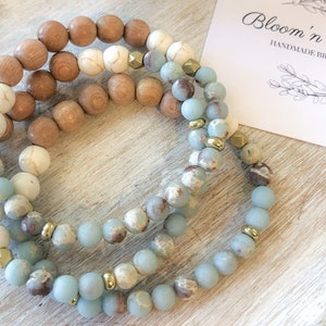 May include: Three handmade bracelets with wooden, white, and blue-grey beads, accented with gold-colored metal spacers. The bracelets are displayed on a light-colored wooden surface. A card with the text "Bloom'n Beads" is in the background.