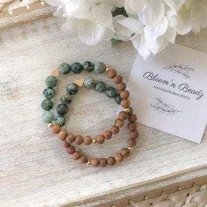 May include: Two bracelets, one with green and brown beads and the other with brown beads, with gold heart charms. The bracelets are on a wooden surface with a white card that says "Bloom 'n Beadz Handmade Bracelets".