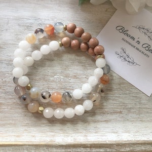 May include: Two bracelets stacked on a wooden tray. The top bracelet is made of light brown wood beads. The bottom bracelet is made of white and light brown agate beads. The text "Bloom 'n Beady Handmade Bracelets" is on a white card.