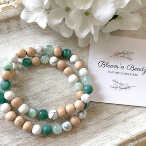 May include: Two handmade bracelets with green, white, and brown beads. The bracelets are stacked on top of each other. The background is a light wood surface. A white card with the text "Bloom'n Beadz Handmade Bracelets" is in the background.