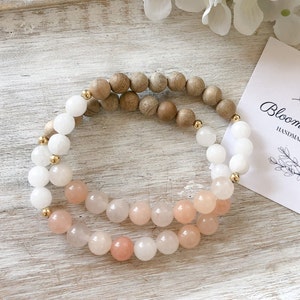 May include: A layered bracelet with white and pink beads, and wooden beads. The bracelet has gold accents.