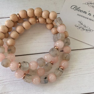 May include: A triple-strand bracelet featuring light pink and gray gemstone beads with gold accents. The bracelet is made with wooden beads and has a "Bloom 'n Beadz Handmade Bracelets" tag.