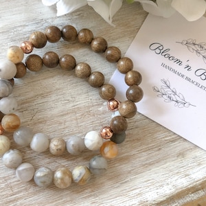 May include: Two bracelets, one with brown wooden beads and the other with light brown, white, and gray beads. The bracelets are stacked on top of each other and have rose gold accents. The bracelets are on a light brown wooden surface. The text "Bloom 'n Beady Handmade Bracelets" is on a pink card in the background.