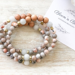 May include: Three layered bracelets with pink, gray, and white beads. The bracelets are made with natural stones and gold accents. The bracelets are displayed on a white wooden surface with a business card that reads "Bloom 'n Beady Handmade Bracelets".
