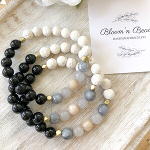 May include: Three handmade bracelets with black, white, and gray beads. The bracelets are stacked on top of each other. The white beads are lava rock, the black beads are onyx, and the gray beads are a mix of light and dark gray. The bracelets are on a wooden surface with a small card that says "Bloom'n Beadz Handmade Bracelets".