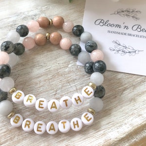 May include: Two bracelets with grey, white, and pink beads, and gold letter beads spelling "breathe". The bracelets are on a wooden surface with a white card that says "Bloom 'n Beadz Handmade Bracelets".