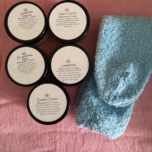 May include: An overhead shot shows five black-lidded jars of Magnesium Cream with white labels and a pair of light blue fuzzy socks on a pink surface. The labels read: Lavender Vanilla, Sunlit Spice, Eucalyptus Mint, Lavendetz, and Sugar Cloud.