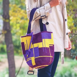 Pode incluir: Uma bolsa tote de crochê roxa e amarela com uma alça longa. A bolsa tem um padrão geométrico e é usada no ombro.