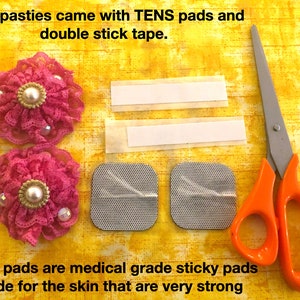 Pu&ograve; includere: Due pasties in tessuto rosa con pizzo e decorazioni di perle, due cuscinetti adesivi TENS, due strisce di nastro biadesivo e un paio di forbici con manici arancioni su uno sfondo di tessuto giallo. "Le tue pasties sono arrivate con cuscinetti TENS e nastro biadesivo." e "I cuscinetti TENS sono cuscinetti adesivi di qualit&agrave; medica realizzati per la pelle che sono molto resistenti" sono scritti in testo nero.