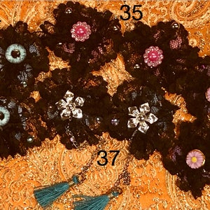 Pu&ograve; includere: Sei applicazioni in pizzo nero a forma di fiore con diversi abbellimenti, tra cui un fiore argentato, una gemma rosa, un occhio blu e una margherita bianca. Le applicazioni sono disposte in fila su un tessuto a motivi dorati. I numeri 34, 35, 36 e 37 sono visibili nell'immagine.