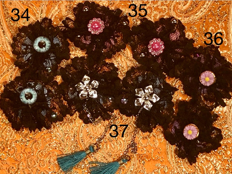 Pu&ograve; includere: Sei applicazioni in pizzo nero a forma di fiore con diversi abbellimenti, tra cui un fiore argentato, una gemma rosa, un occhio blu e una margherita bianca. Le applicazioni sono disposte in fila su un tessuto a motivi dorati. I numeri 34, 35, 36 e 37 sono visibili nell'immagine.