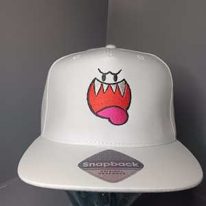 May include: A white snapback cap featuring an embroidered graphic of a stylised ghost character with a wide red mouth, sharp white teeth, and a pink tongue. The cap includes a diamond-shaped sticker on the flat brim that reads Snapback.