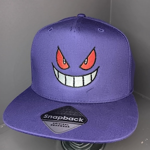 May include: A purple snapback cap featuring an embroidered graphic of a mischievous face with sharp red eyes and a wide, toothy grin. A black sticker on the flat brim displays the text Snapback Original Headwear in white and silver lettering.